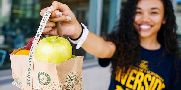 Basic Needs content module thumbnail UCR student holds up bag of free fruit.